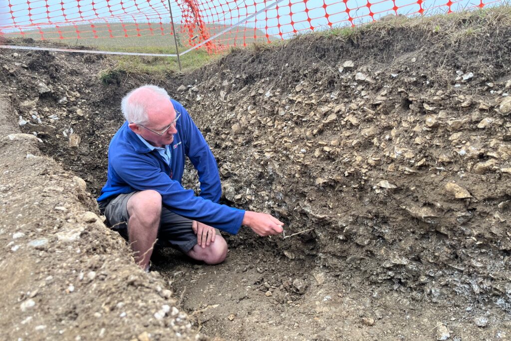 A man in a blue fleece and shorts kneels in a trench. He is holding a trowel and using it to remove earth from the side of the trench. 