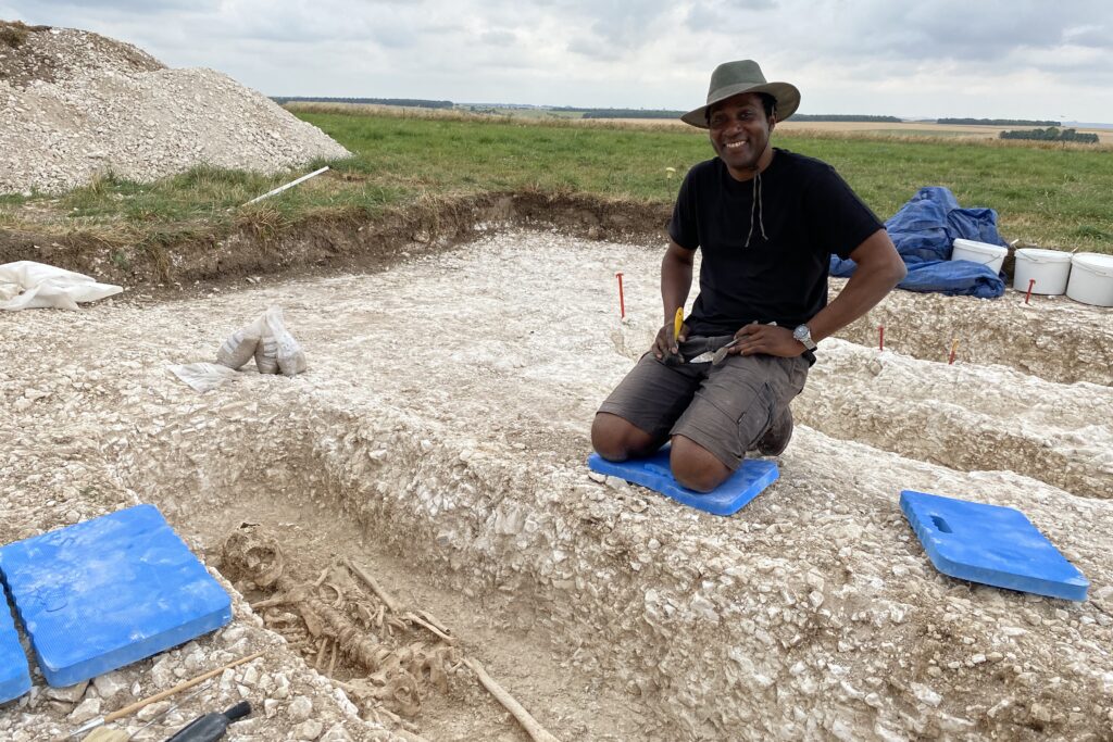 A man in a t-shirt, trousers and a hat kneels next to an excavated grave, containing a skeleton, and smiles at the camera