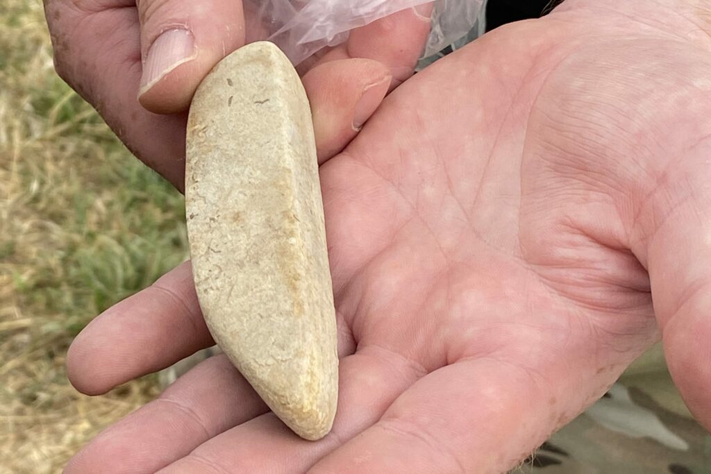 Hands holding a light-coloured stone, approximately two inches in length. It is broadly rectangular, with one of the long sides appearing flat while the other is curved.
