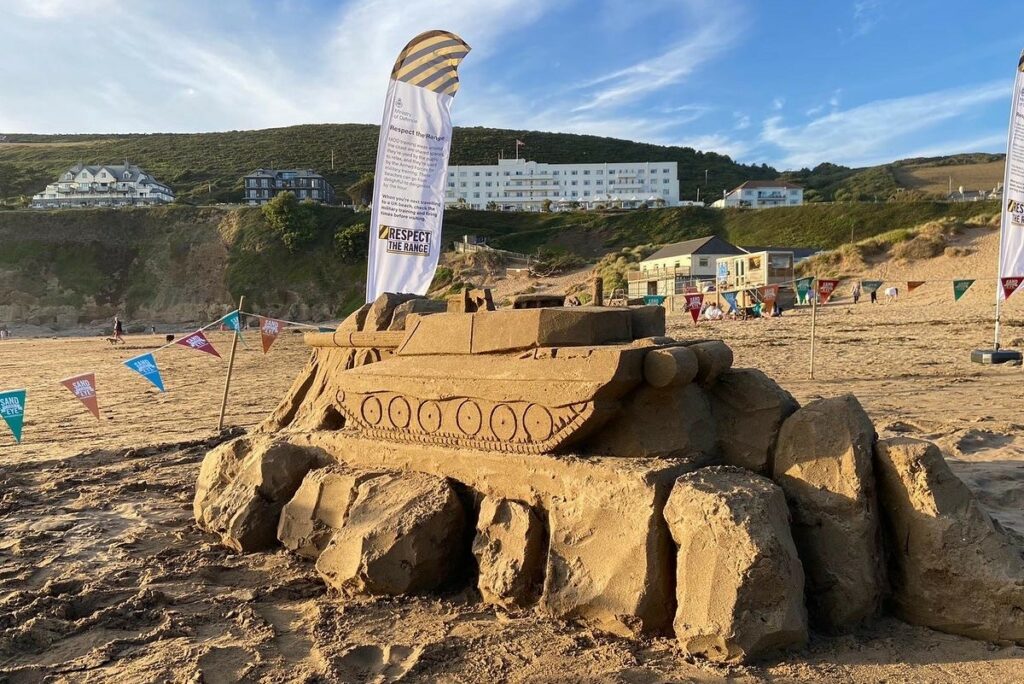 A sand sculpture of a Type 2 Challenger tank on a beach. Behind the sculpture is a flag displaying the 'Respect the Range' slogan.