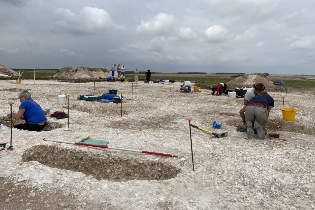 Landscape view of what appears to be a chalky area with several graves excavated. There are a number of people kneeling over different graves with more standing in the background.
