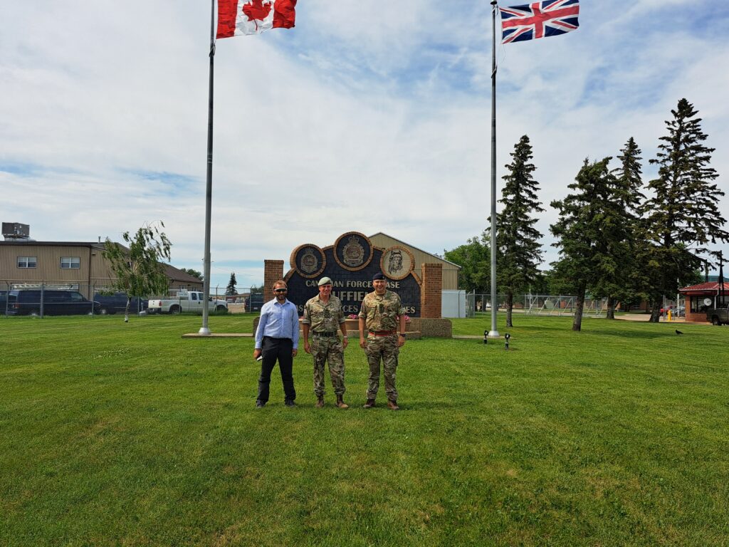 Three men, two in military uniform and one in civilian clothing, are stood in a grassy field with buildings to either side. Behind them is a large plaque reading 'Canadian Forces Base Suffield.' Next to the plaque are a pair of flags, one displaying the Canadian flag and the other displaying the flag of Great Britain.