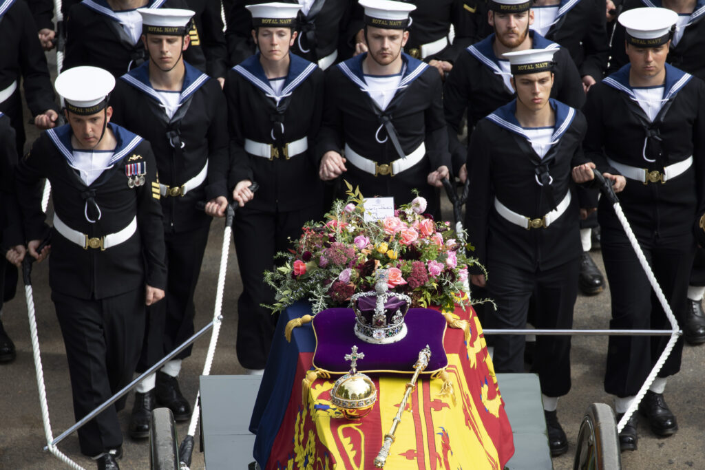 HM The Queen's coffin, topped by the Royal Standard, Crown Jewels and a large bouquet of flowers, with a number of Royal Navy sailors holding ropes behind it. 