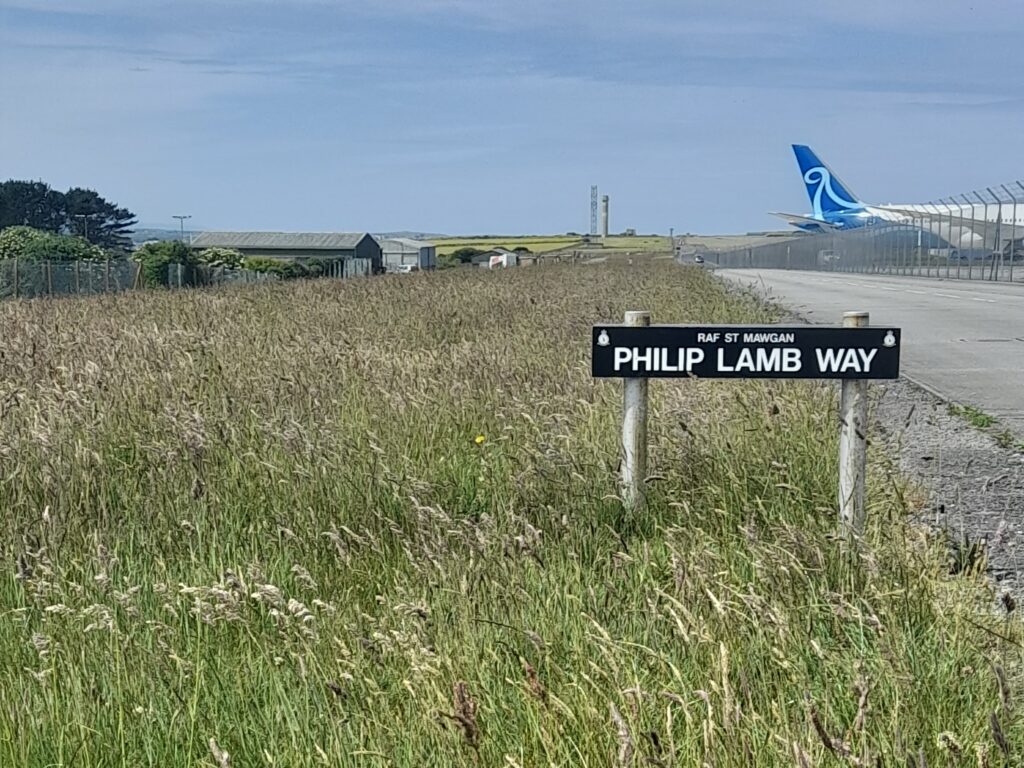 A photo showing a field of tall grass, with buildings to the left and an airport to the right. In the foreground is a sign reading 'RAF St Mawgan: Phil Lamb Way'
