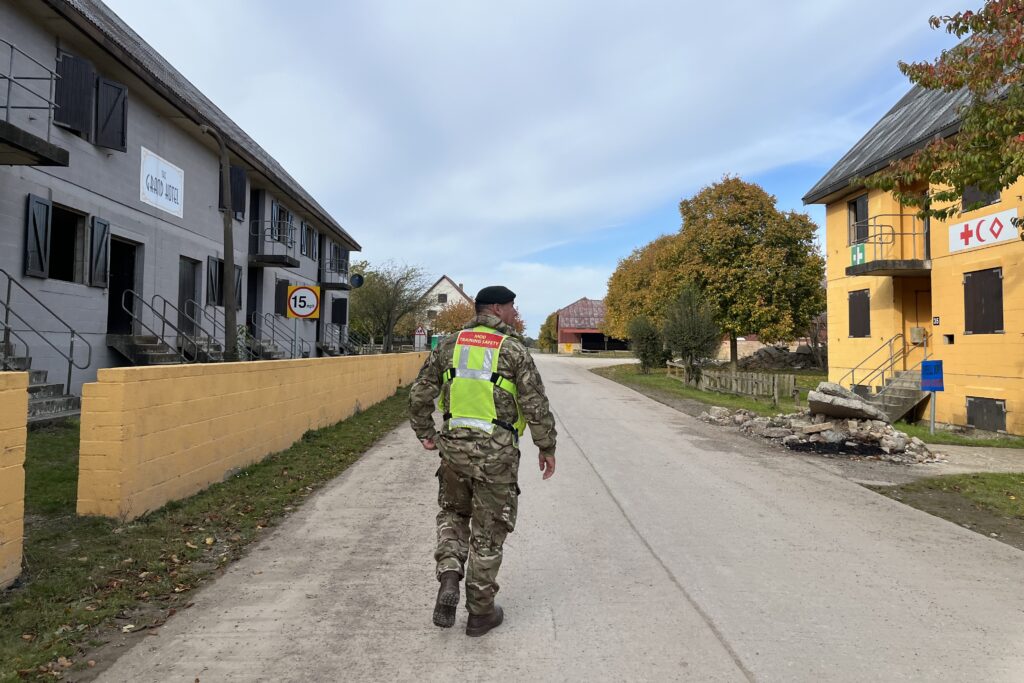 A man wearing Army camouflage uniform under a high vis vest walks down a street in Copehill Down the urban training facility. There are buildings to either side.