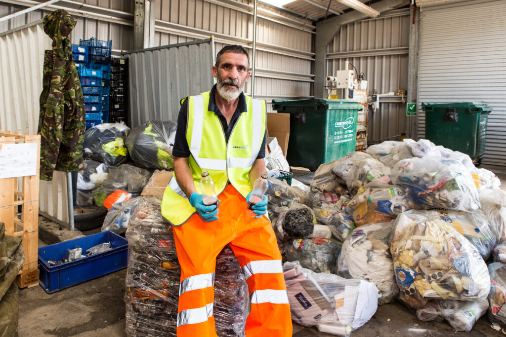 A man in hi vis vest and trousers sits on top of a clear bag full of waste. He wears blue gloves and in his hands are two plastic bottles.