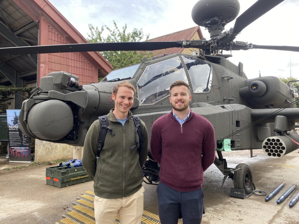Two white males in smart-casual clothing, stood in front of an Apache helicopter.