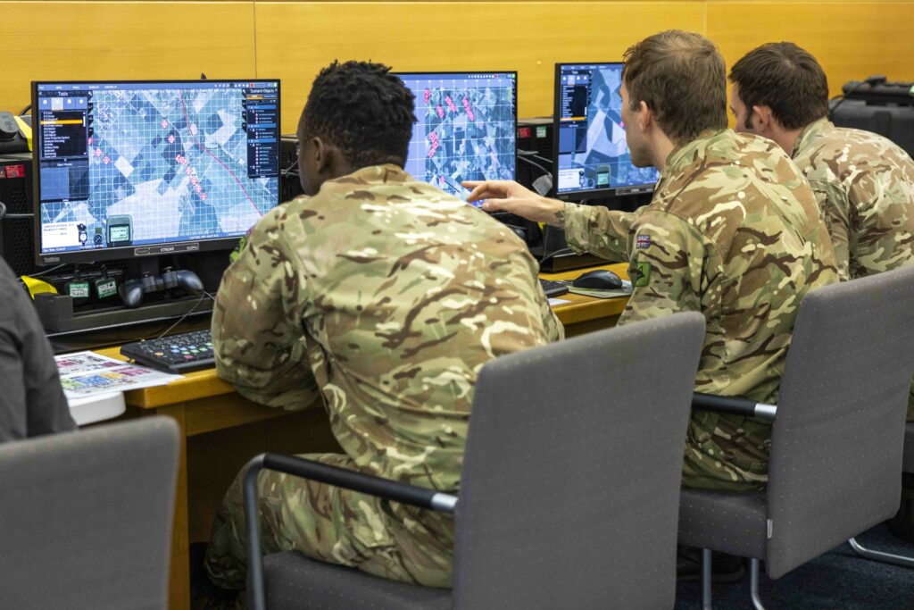 Three men in military uniform sit in a room with yellow walls. Each man sits infront of a computer.