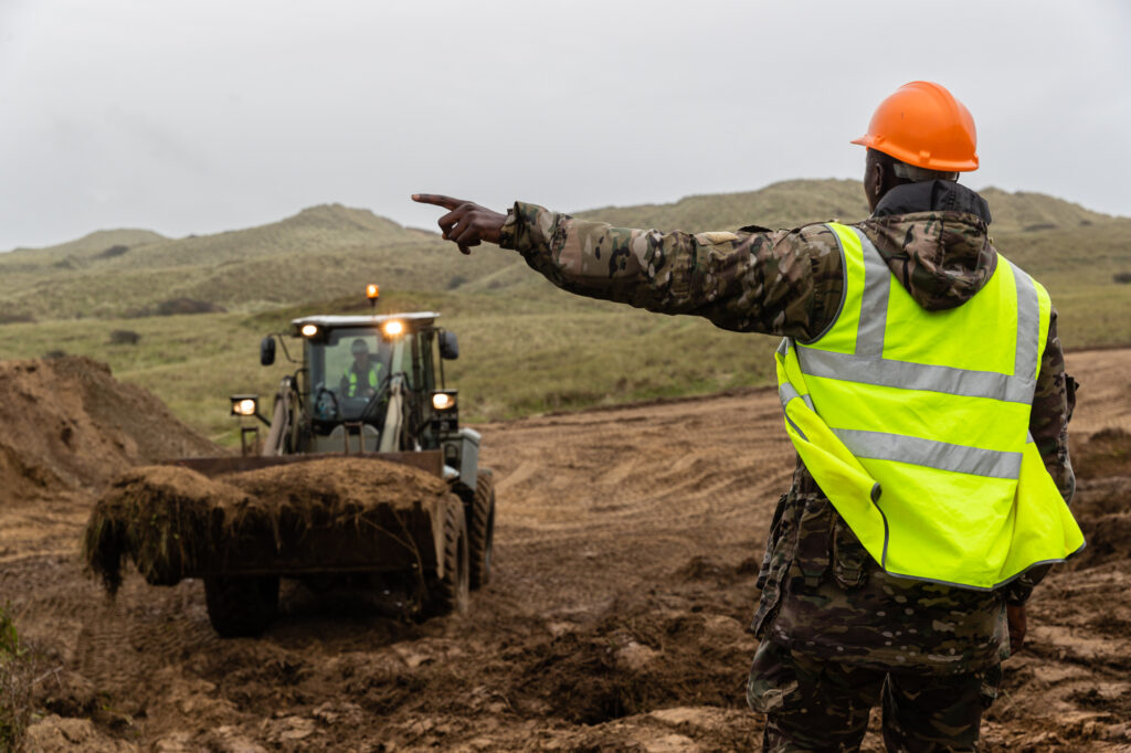 In the foreground a man is wearing military camouflage clothing, a high vis yellow vest and an orange hard hat. He directs a military digger which is lifting a pile of soil
