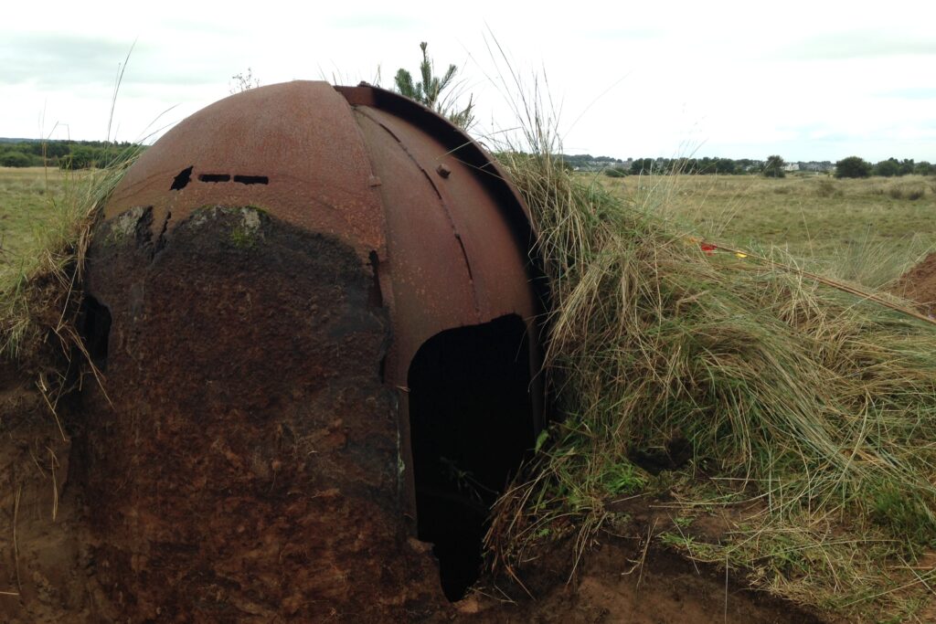 A rusted metal, dome-shaped object with a square shaped opening in its front, surrounded by mud and grass.