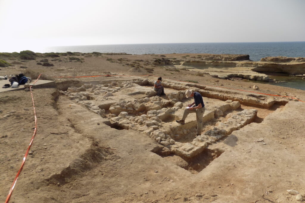An excavation site with various layers of rocks and sand into which trenches have been dug. The site is bordered by the sea in the background. Two archaeologists, one ale and one female, are stood near the centre of the image, writing notes.
