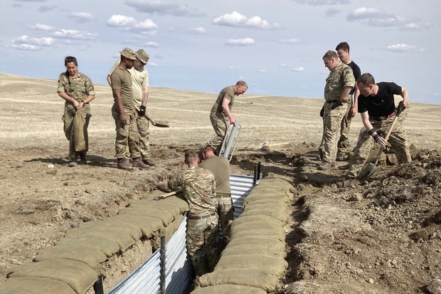 A number of personnel in military uniform are stood above and within a section of trench dug into the ground. They are carrying shovels and other equipment.
