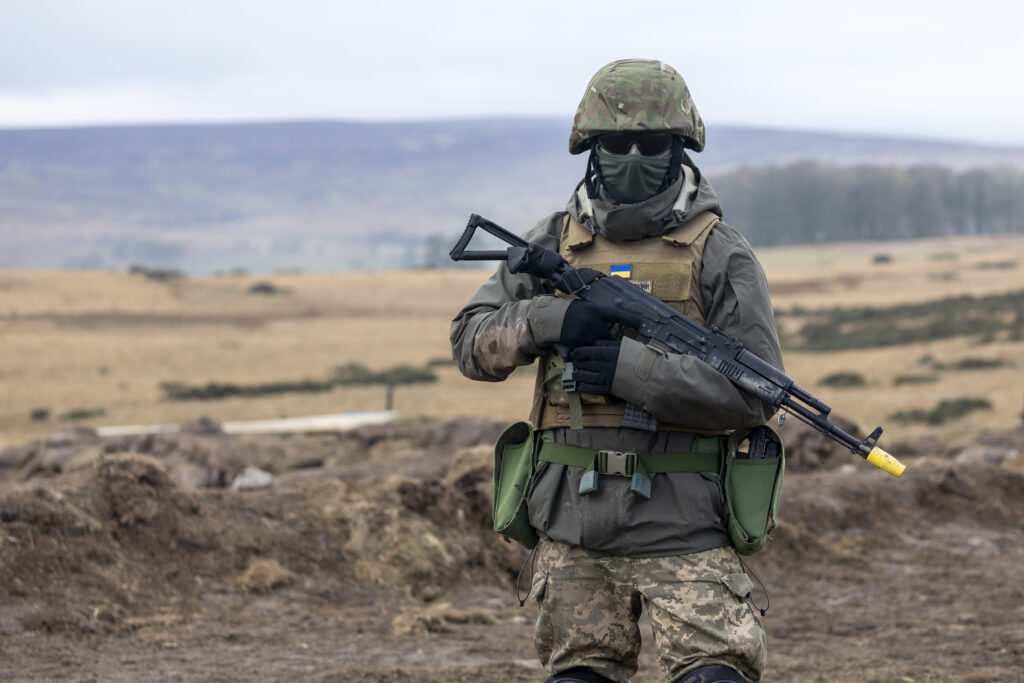 A Ukrainian recruit faces the camera in uniform and holding his gun in a relaxed position in his arms. His face is entirely covered by sunglasses and a mask or balaclava. There is an open expanse of ground behind him. 