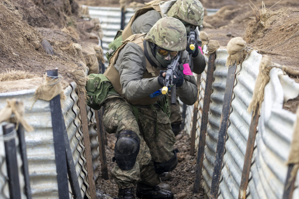 Ukrainian recruits move at a slight crouch towards the camera through a trench system lined with corrugated metal.