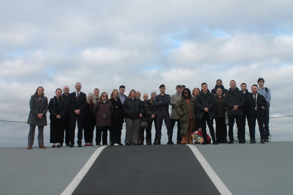 A group of about 25 people stand looking down at the camera from the top of a grey ramp. The ramp is mid grey on either side and dark grey in the middle.