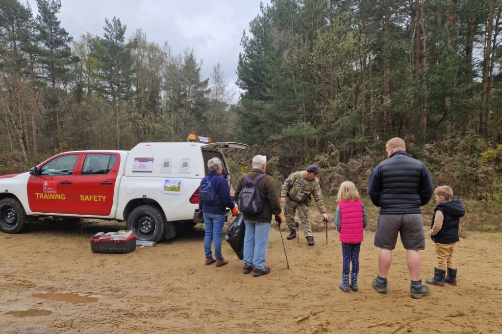 A man in a camouflage military uniform speaking to a small group of adults and children. They are stood next to a red and white 4x4 vehicle with 'Training Safety' written on its side