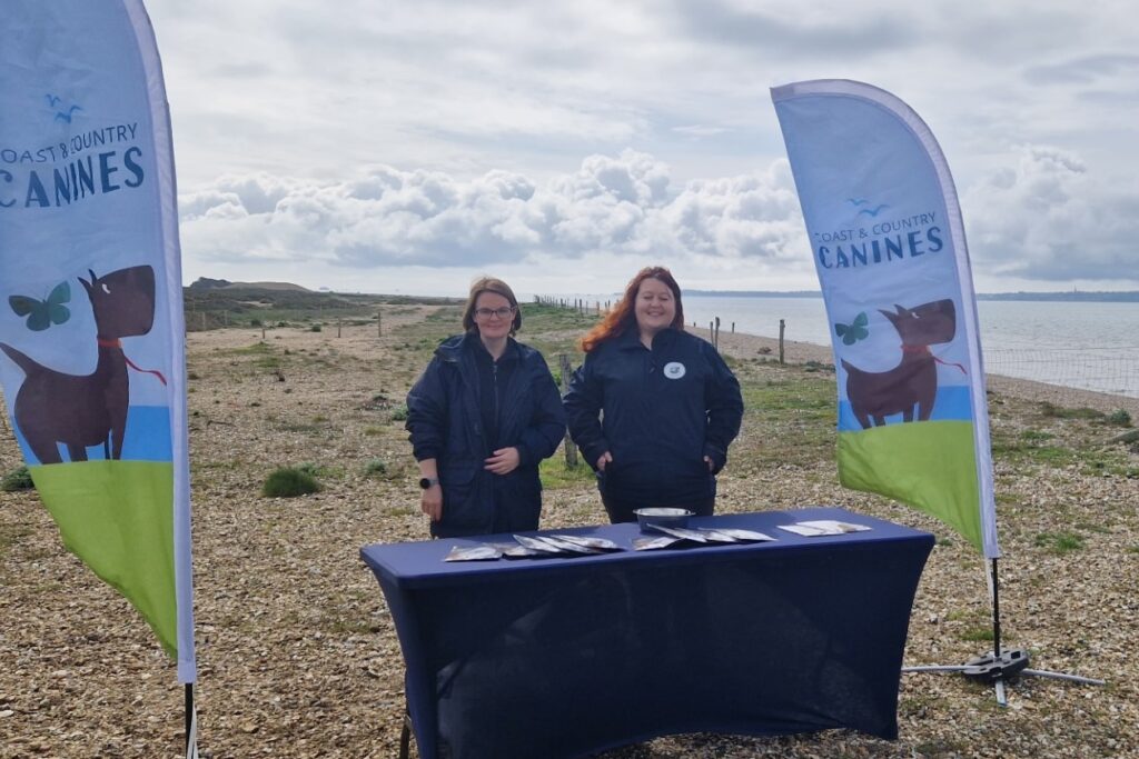 A pair of women in navy blue uniforms stood behind a table on a beach, with flags to their left and right reading 'Coast and Country Canines'.