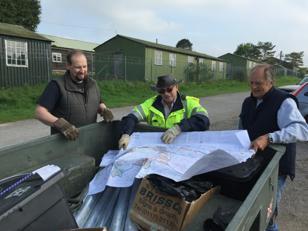 Three men examine a map unfurled in the back of a pick-up truck. Behind them is a row of green, single storey military buildings bordered by a fence and a tarmac road.
