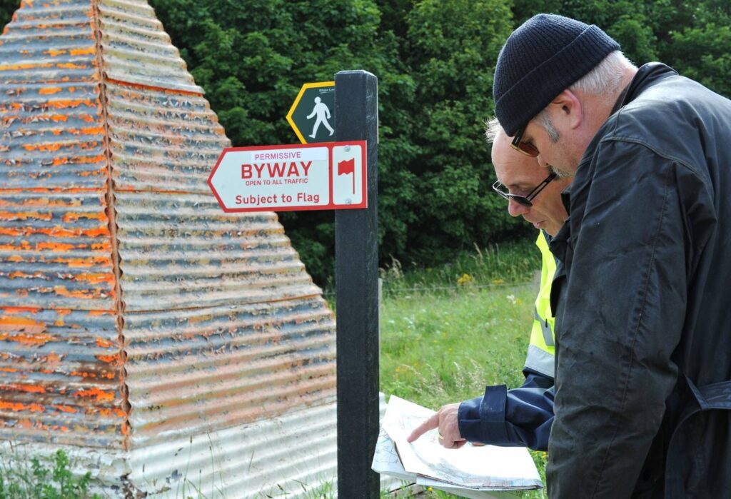 Two men, one wearing a dark leather jacket and the other wearing a high-visibility jacket, examine a folded section of a map. Next to them, at head-height, is a sign which reads: ‘Permissive Byway / Open to all traffic / Subject to flag’