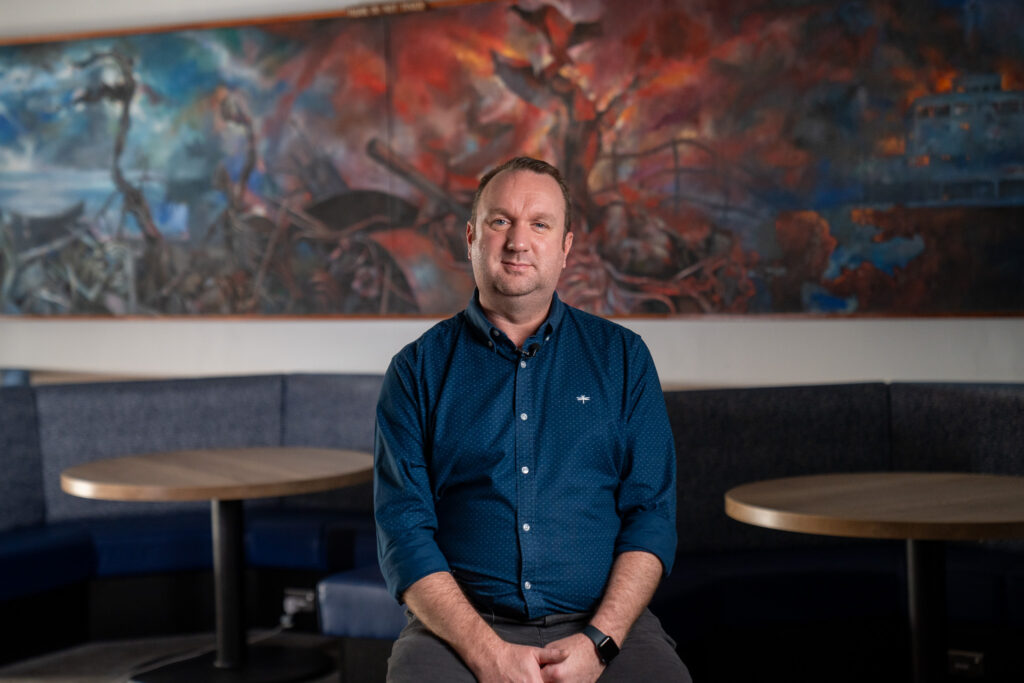 A DIO employee wearing a blue shirt is seen sitting in front of a striking painting depicting the Falklands War. The abstract image features blues and oranges as it show the Bluff Cove air attacks in June 1982.