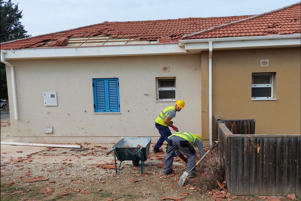 Two workmen in high vis and helmets, with a wheelbarrow, clear up the outside of a Service Family Accommodation property. There is clearly some damage to the roof with missing tiles.