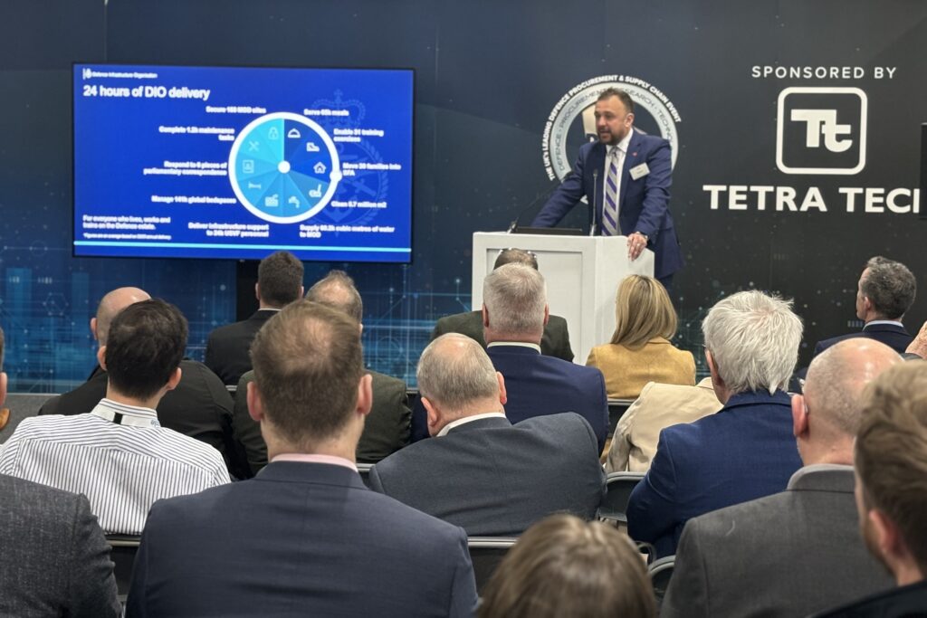 Lee Taylor presenting his speech at the 2024 DPRTE event, to an audience of numerous seated people. He stands behind a lectern and has a screen to his right with a pie chart saying '24 hours of DIO delivery'. The wall behind him reads 'Sponsored by Tetra Tech'. 