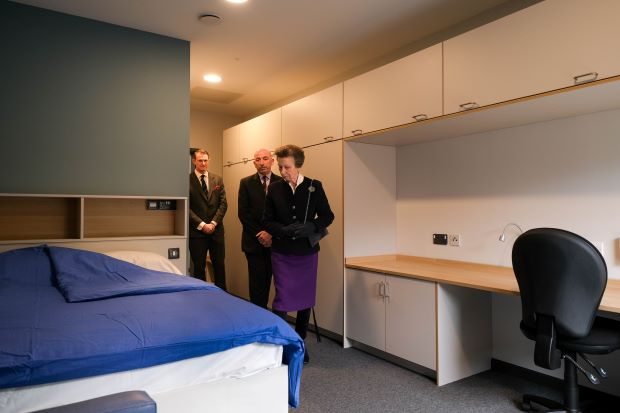 The Princess Royal is in a bedroom in a military accommodation block. In the room is a white desk with a black office chair and a bed against the wall with a blue blanket covering it.
