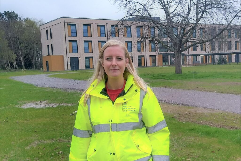 A woman wearing a high visibility jacket stands in front of a newly completed accommodation block. There is grassland around.
