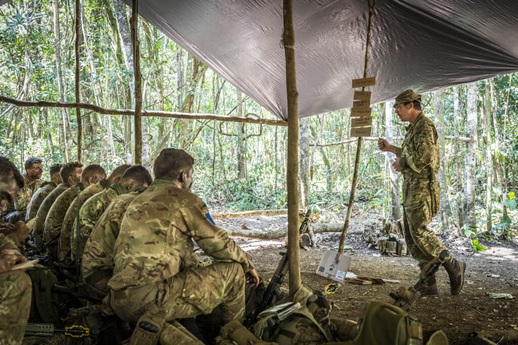A group of soldiers sit in two rows, listening to another soldier who stands in front of them. They are in the jungle covered by a rudimentary shelter of a tarp held up by branches. 
