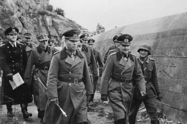 A black and white picture of members of the Nazi Party in military uniform walking next to a concrete wall. 