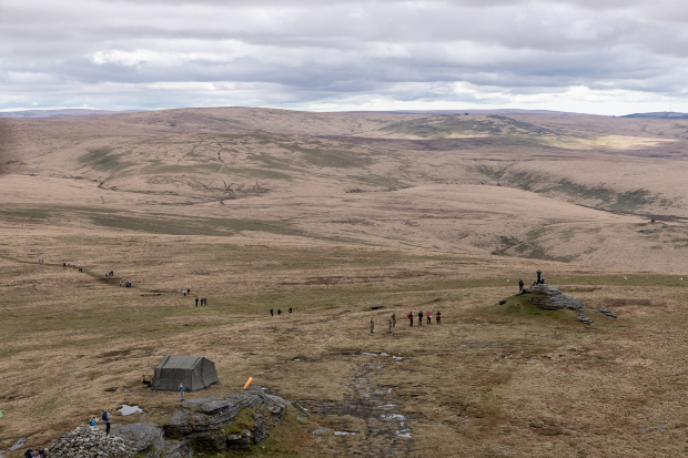 Aerial photo showing the rugged terrain at Dartmoor as a line of walkers heads uphill to a tor with a checkpoint tent. 