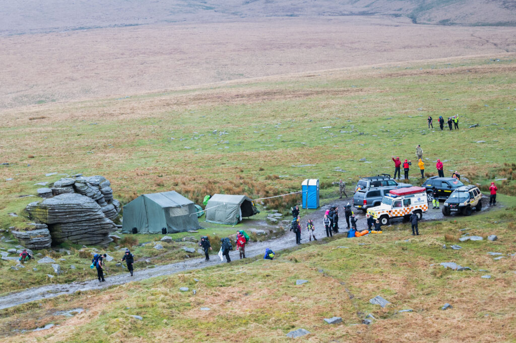 Aerial photo of a group of people standing on a track in an open, featureless area of moorland. There are several vehicles and a few large tents. 