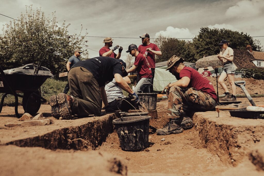 A group of people are kneeling by a dig out in the ground. A black bucket is seen and the workers are looking at archaeology site.