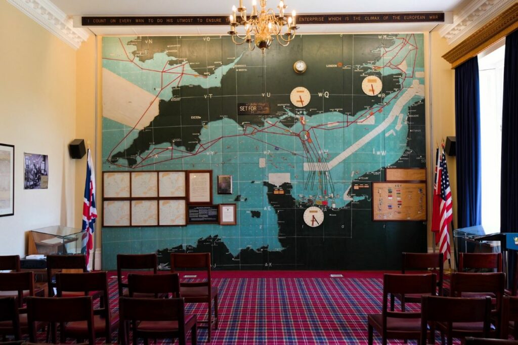 A room with an old blue map showing the south coast of England and the northern coast of France. This map was used to plot the D-Day landings. Lines are seen on the map to show routes to be taken by Royal Navy shops, and various clocks showing different times are also seen.