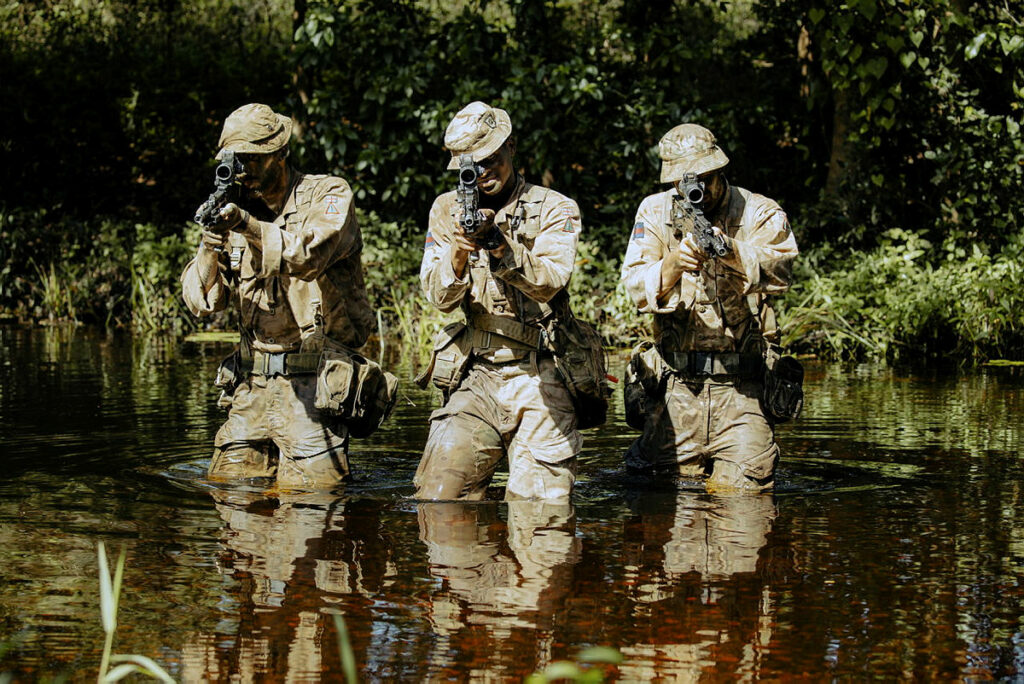 Three soldiers wading through a river up to their thighs. They are holding rifles pointing towards the camera.