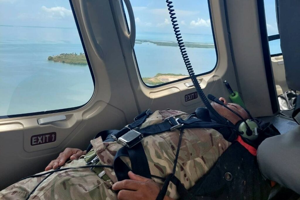 A member of the training team, wearing military uniform, is strapped horizontally into a flying helicopter. The photo is taken from inside the helicopter and the sea and some islands are visible through the windows.