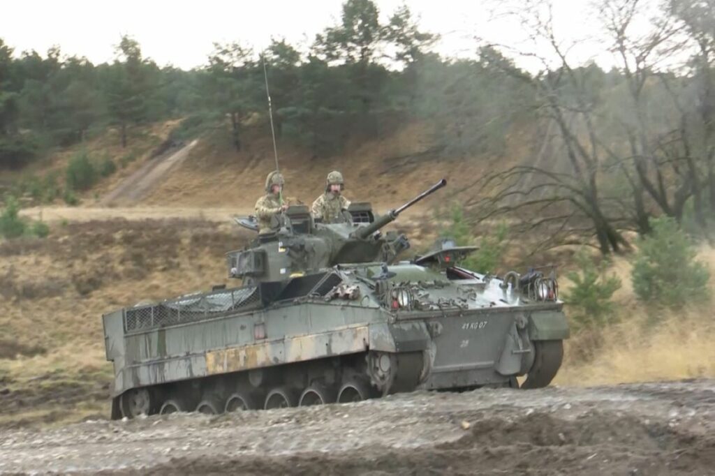 Two soldiers in uniform sit on top of a military vehicle on a training area in Germany. 