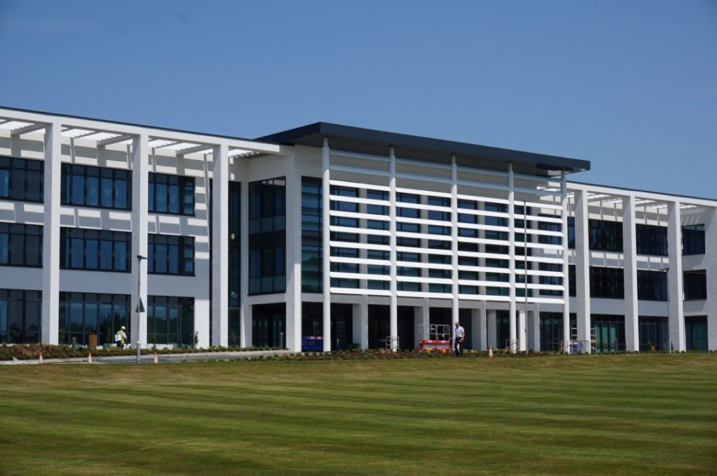 The College is a long modern looking white building with white pillars infront of dark blue modern looking windows. There is grass infront of the building.