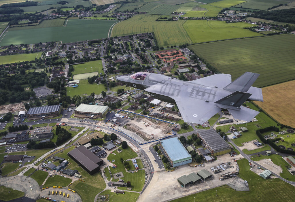 A grey F-35B Lightning aircraft flys over the RAF Marham station. There is a range o hangars and buildings beneath the plane with lots of greenland around the station