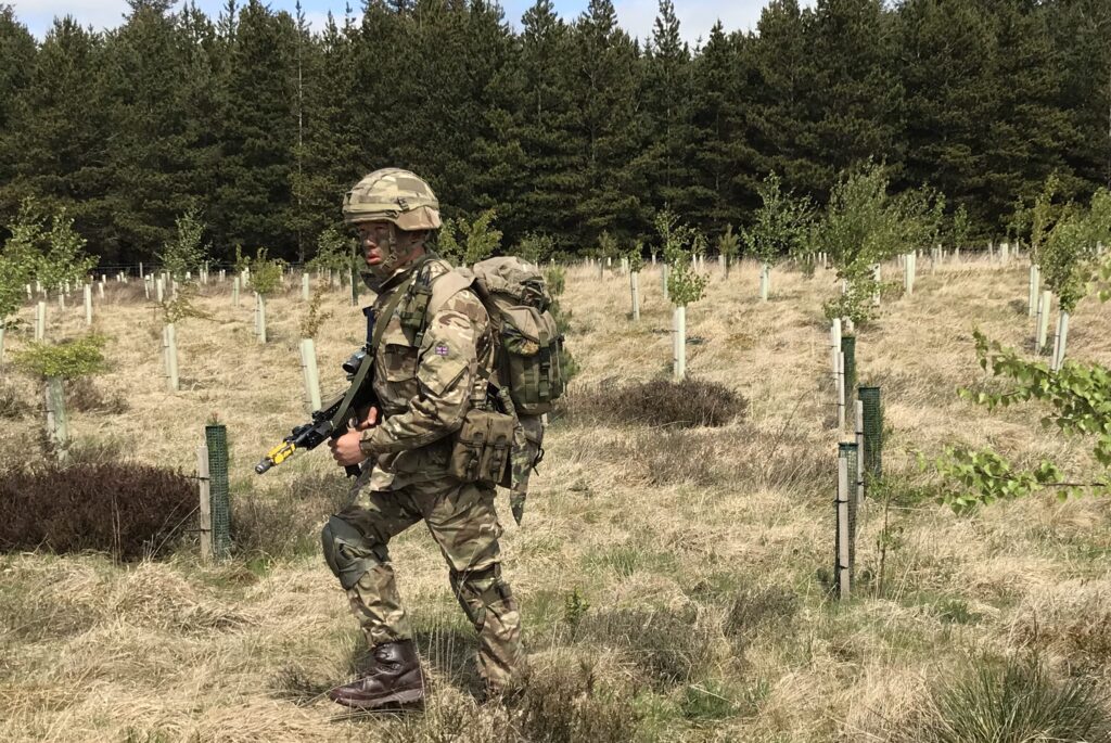 A Gurkha recruit, in full uniform including helmet and body armour, and carrying his weapon and a bergen, moves through an area of growing trees with a mature copse in the background. 