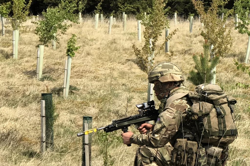 A Gurkha trainee, wearing full uniform including helmet and body armour, and carrying his weapon and a bergen, kneels in an area of young, growing trees. A mature wood is in the background. [Crown Copyright/MOD2018]