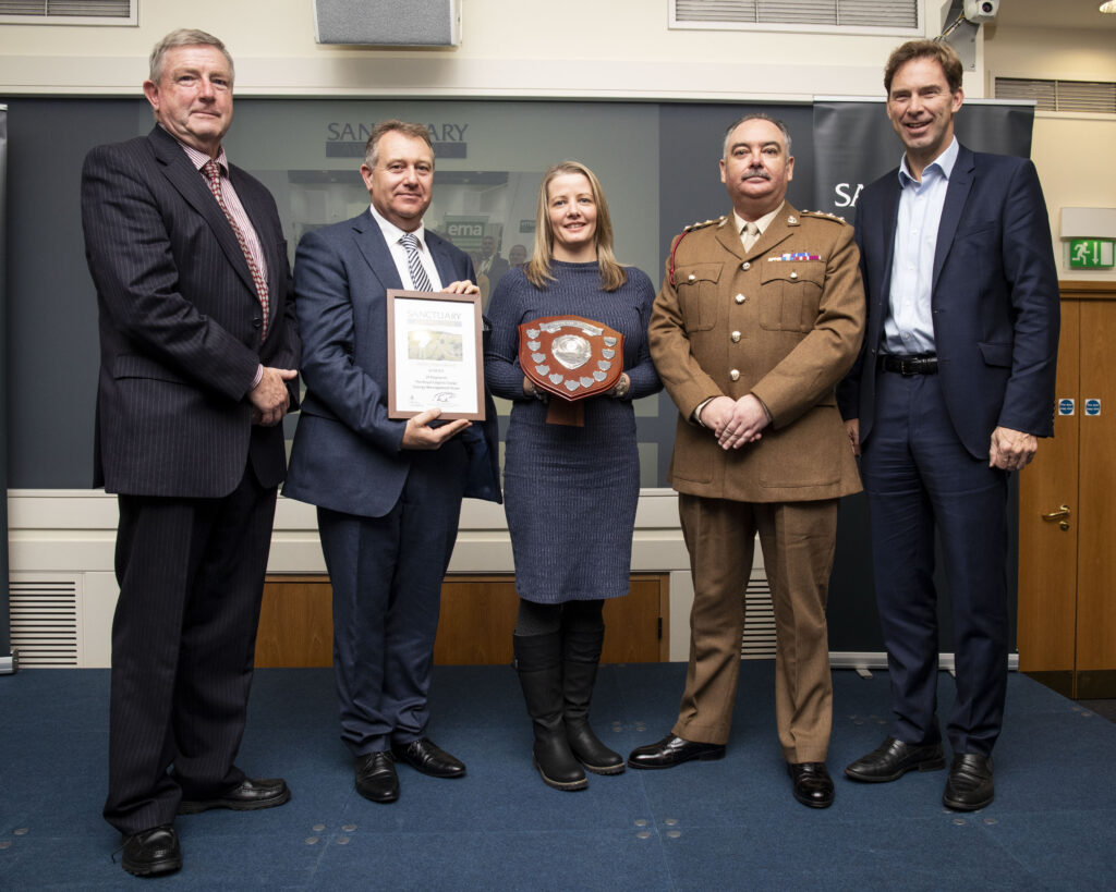 Pictured are five individuals. On the left is a white male i a black suit, pink shirt and tie, next to him is another white male in a dark grey suit, white shirt and grey tie holding up a square framed award, next to him is a white female with short blonde hair wearing a grey dress and long black boots holding up a brown shield shape award with mini silver shields around it. On the right of her is a white male dressed in brown military wear and next to him was then Minister for Defence People and Veterans Tobias Ellwood dressed in a black suit and white shirt. 