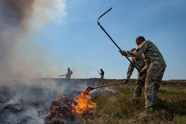 In the foreground, a soldier from 4 SCOTS holds a long implement in the air, in the middle of using it to beat down flames in the centre of the image. In the background are other soldiers.