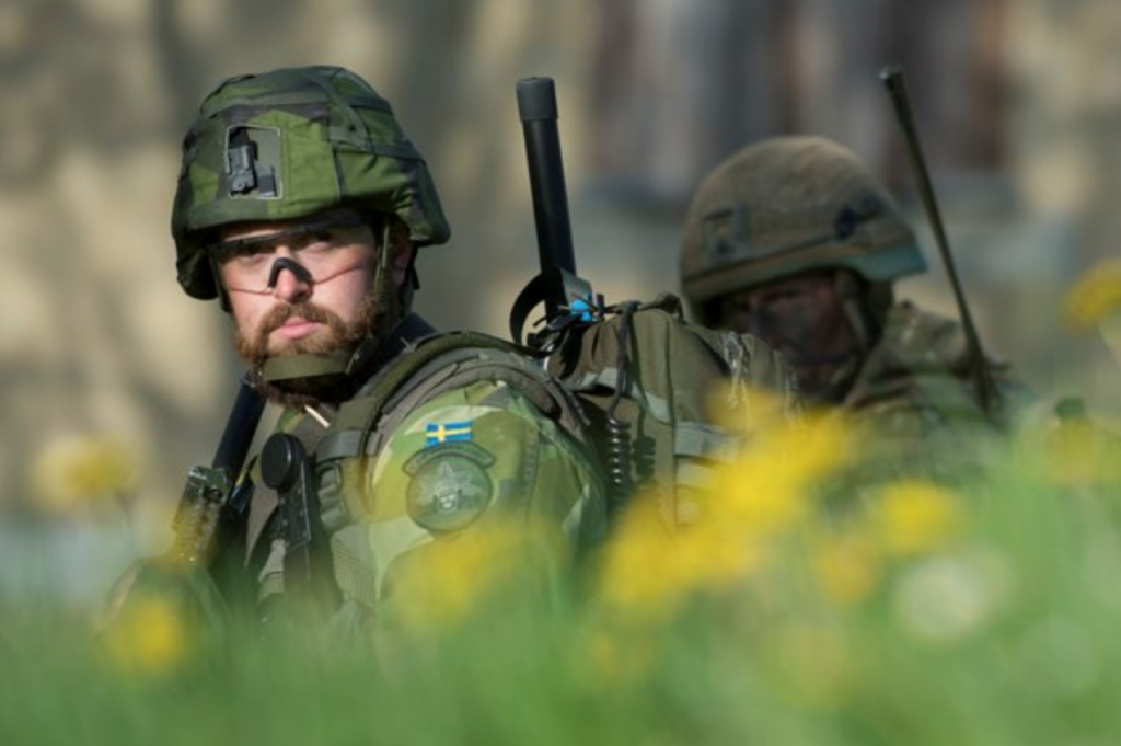 A Swedish soldier, in uniform and standing in profile, is looking towards the camera, with a British soldier behind him.