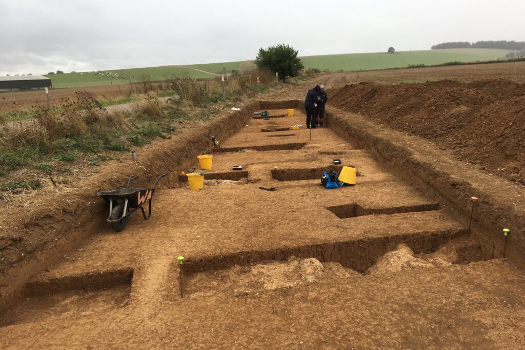 A wide, shallow trench with a number of deeper trenches within it. There is a wheelbarrow and several buckets and two people at the far end conversing.