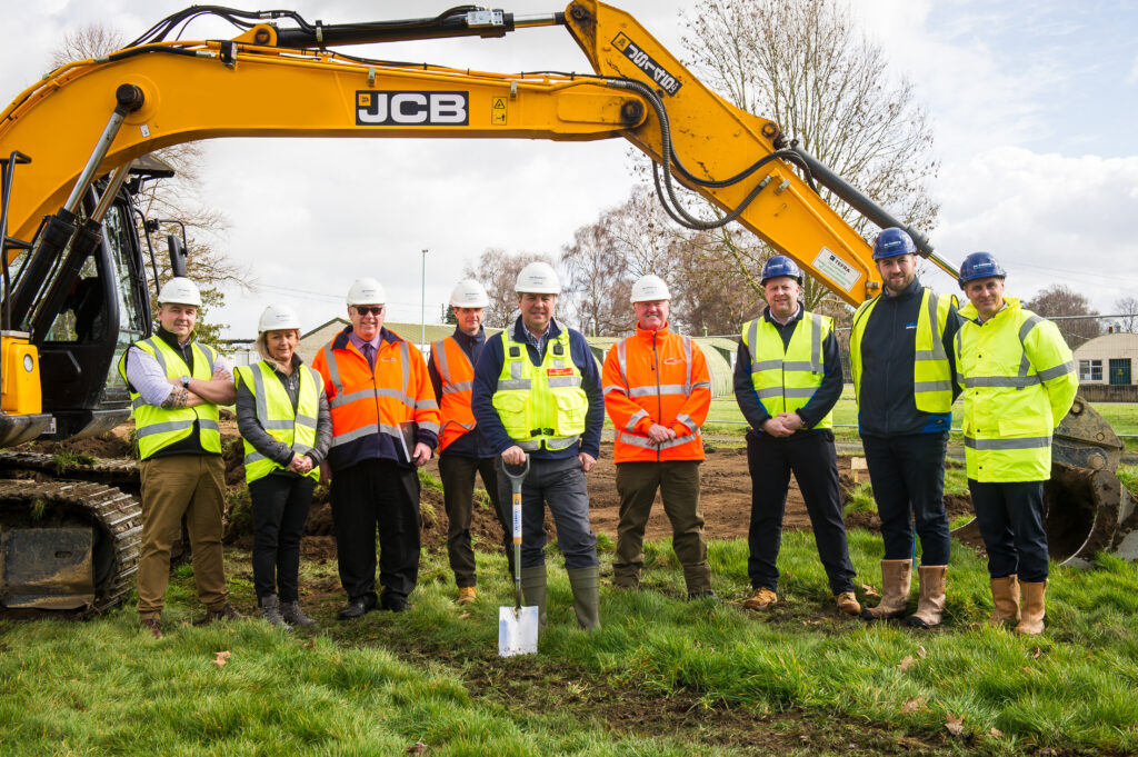 Representatives of DIO, Landmarc and Pave Aways Ltd attend the ground breaking ceremony. They are all dressed in helmets and high vis jackets and one holds a spade. They are standing on grass in front of a building. 