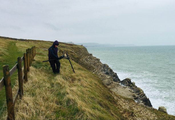 Keri Thomas, from the Environmental and Ordnance Liability team at DIO carrying out a check with a piece of equipment on the cliff edge of the South West Coast Path Trail in Dorset. She is standing on grass at the edge of the cliff with a sea view.