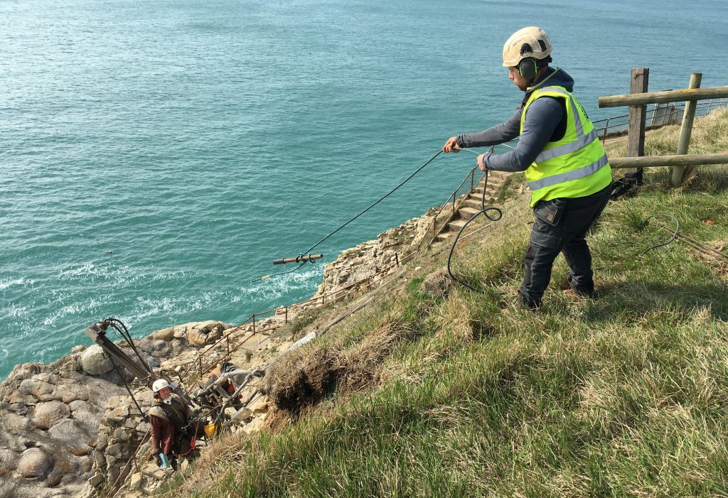 A member of the Vertical Technologies team, dressed in a helmet and reflective jacket swings a rope down the cliff to a colleague.