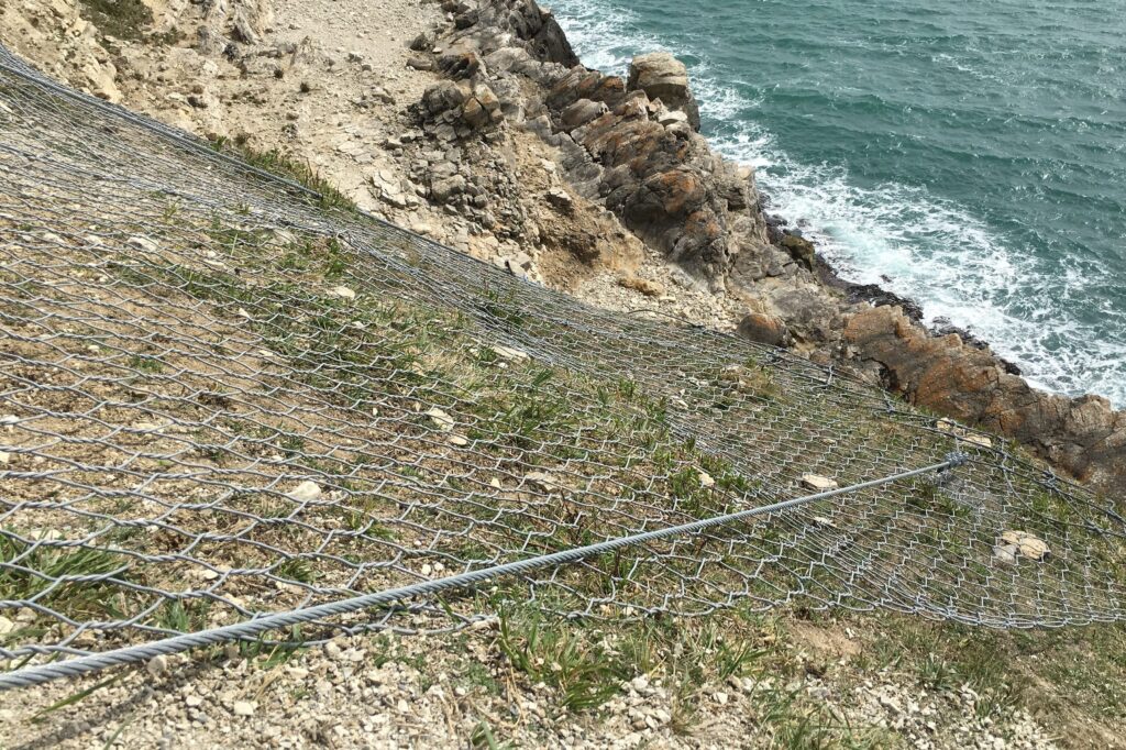 The image shows metal netting over the rocks. It is taken from the top of a steep slope looking down towards the sea. 