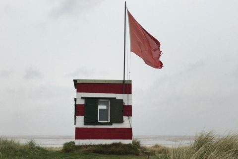 In the foreground is a shallow grassy slope and behind appears to be the beach. In the centre of the image is a small building with red and white horizontal stripes and a large red flag.